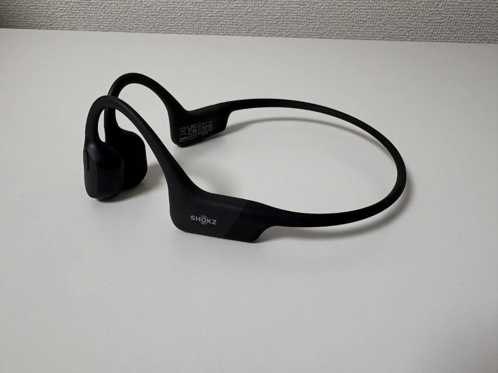 2年使った Shokz OpenRun。白い机に置いた本体を斜め上から見たところ。黒いネックバンドとイヤーフック、バンドの外側に SHOKZ のロゴ