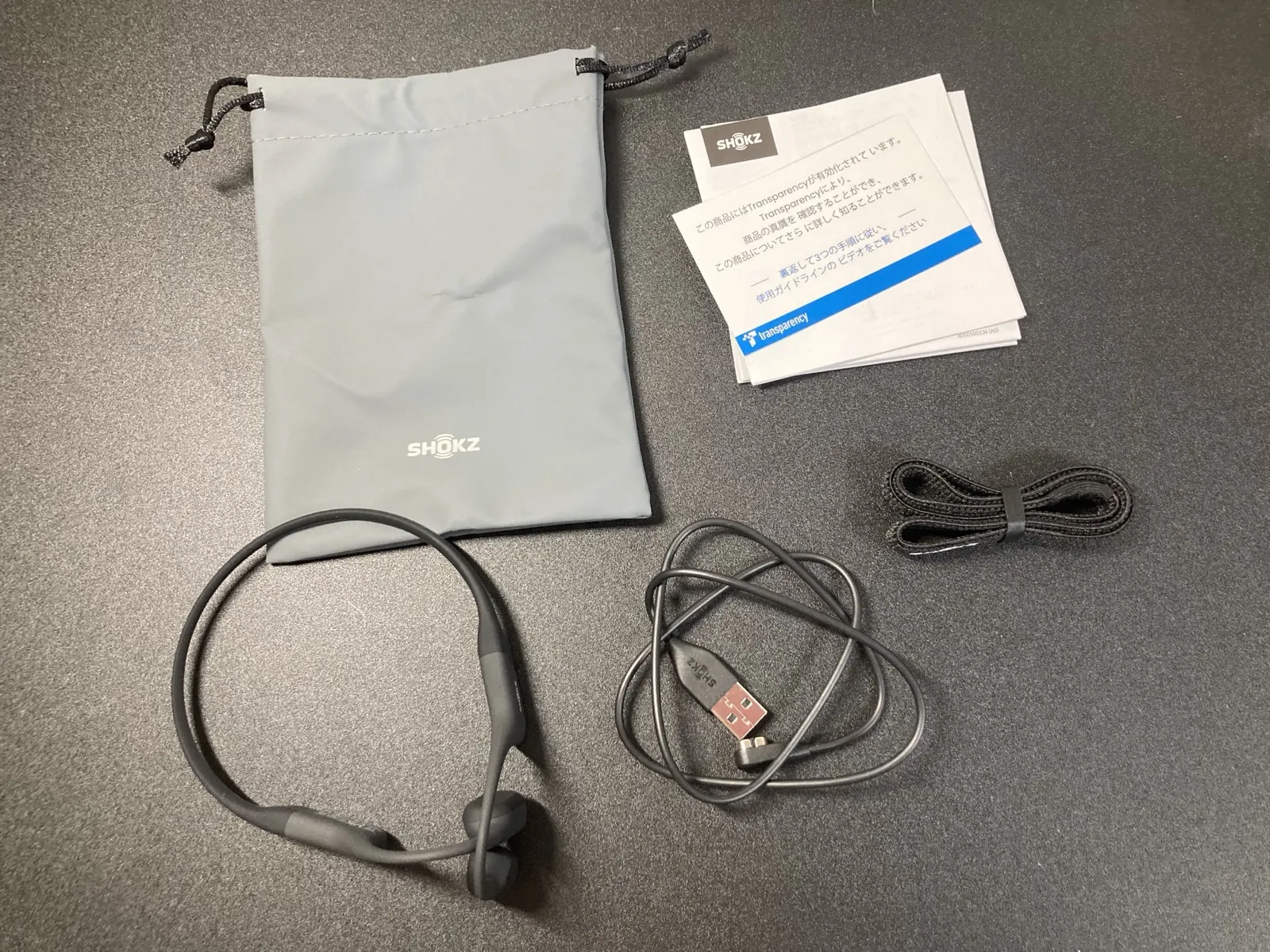 同梱物一式。Shokz のロゴ入りポーチ、日本語の説明書、本体、マグネット式の充電ケーブル、予備のストラップ